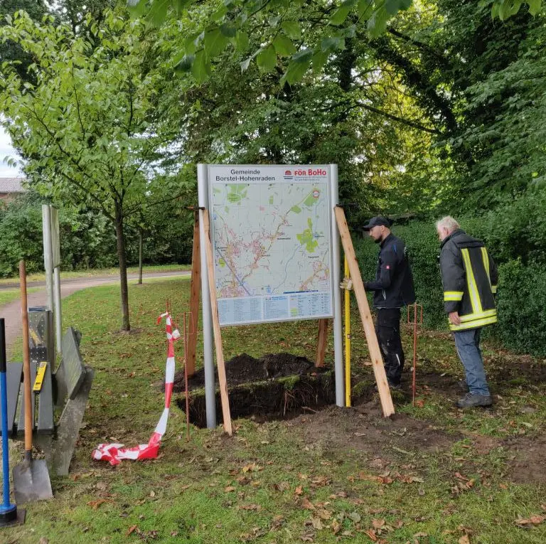 Aufstellen der ersten Info-Tafel: Tafel mit Dorfplan und Helfer an der Baugrube